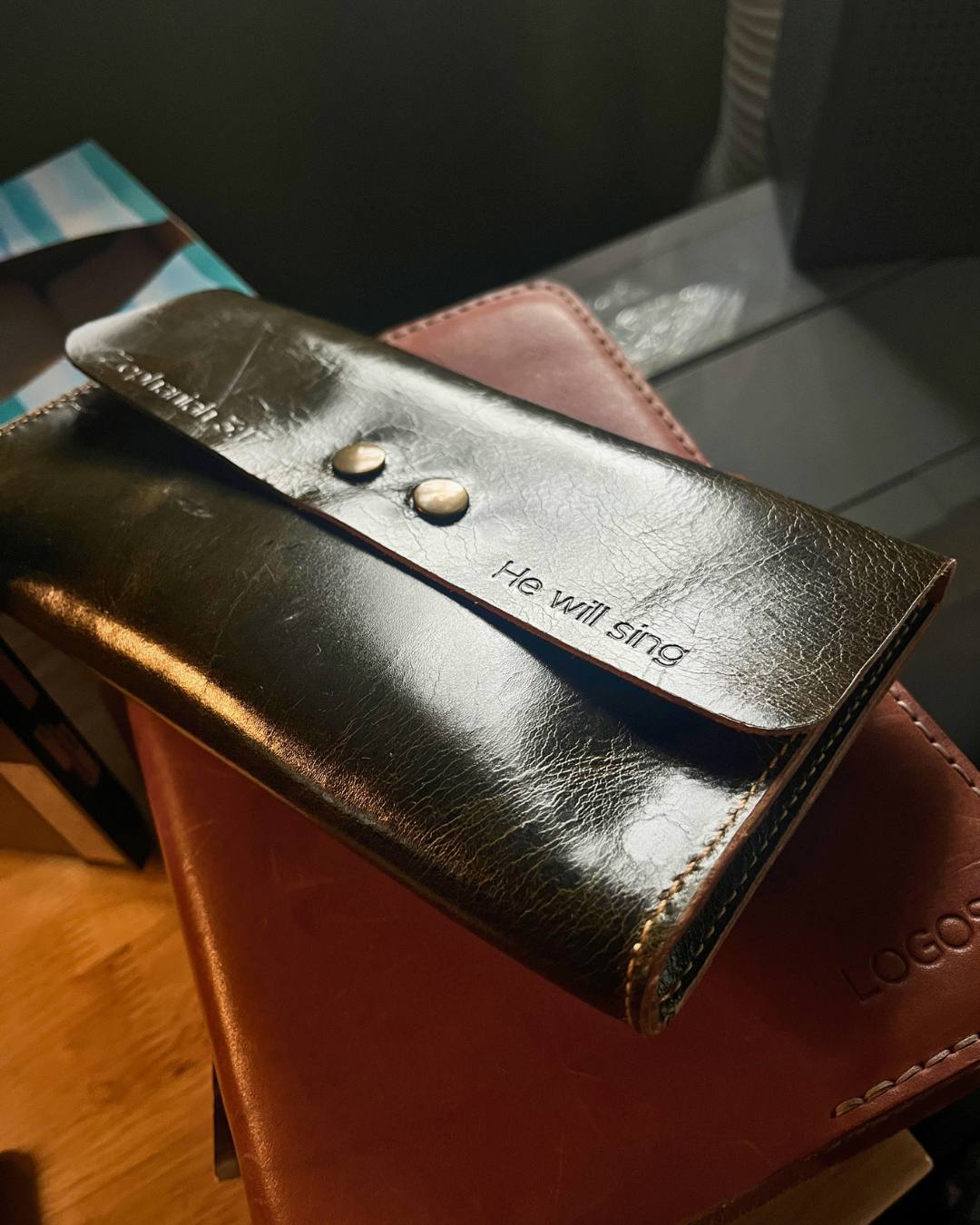 Green leather wallet with 'He will sing' engraving on a wooden surface