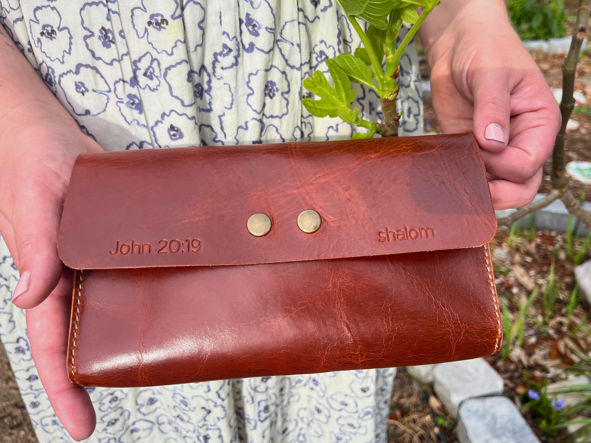 Brown leather wallet held by hands with 'John 2019' and 'shalom' engraved, outdoor background.